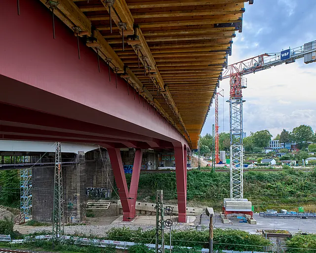 Die Unterseite einer Brücke auf roten V-Förmigen Stahlpfeilern über Bahngleisen.