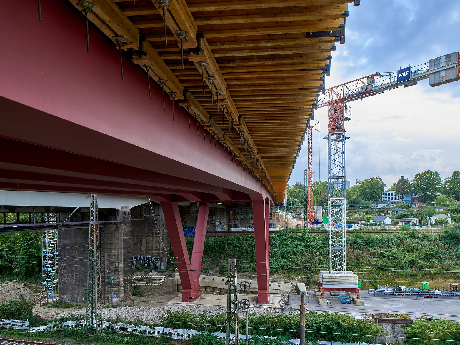 Die Unterseite einer Brücke auf roten V-Förmigen Stahlpfeilern über Bahngleisen.
