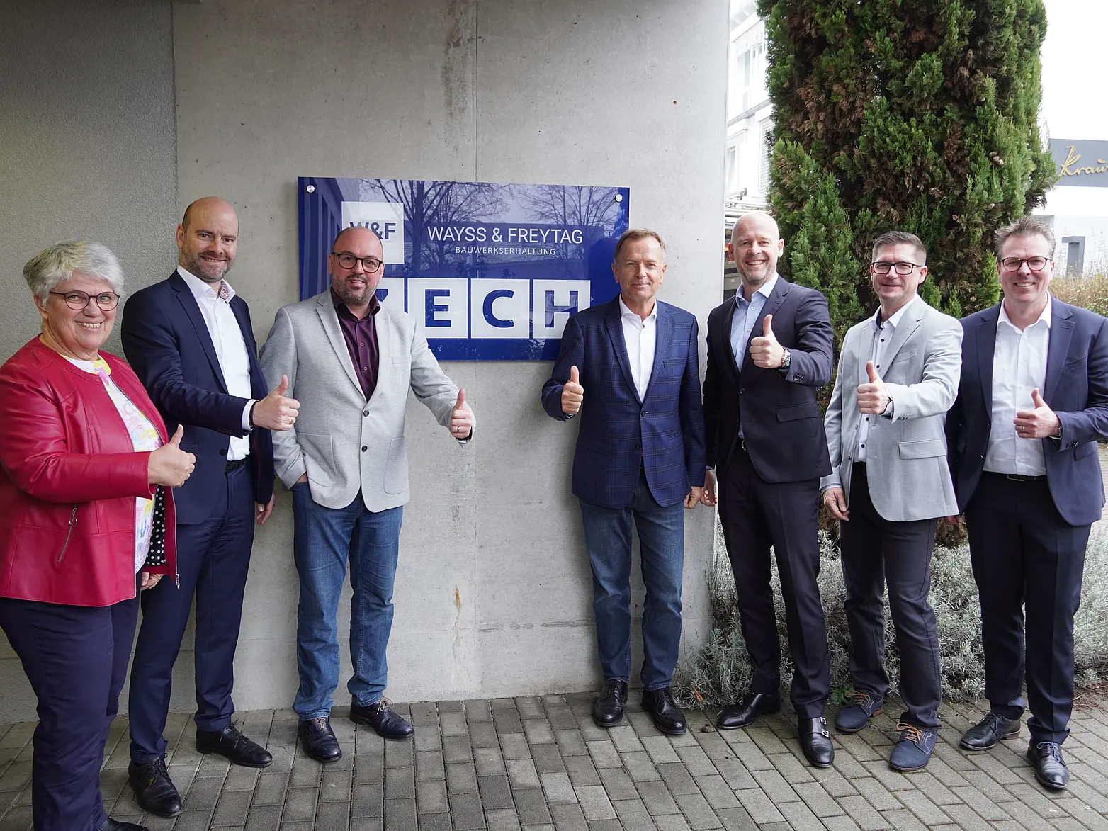 Personen stehen vor einem Unternehmenslogo und zeigen Daumen hoch.