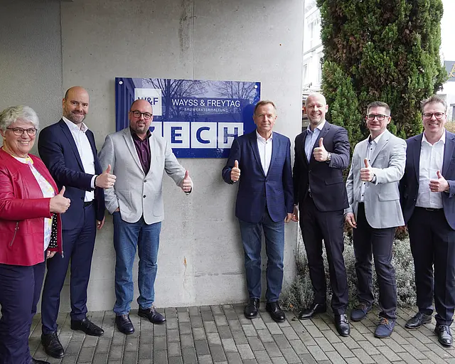 Personen stehen vor einem Unternehmenslogo und zeigen Daumen hoch.