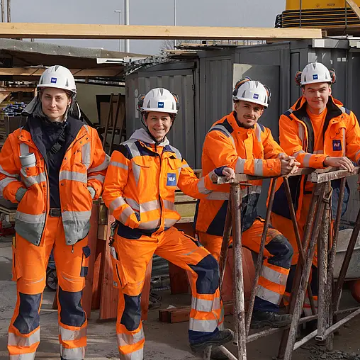 Vier Auszubildende sind auf einer Baustelle in orangener Sicherheitskleidung.