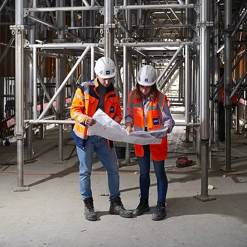 Zwei Mitarbeiter stehen zusammen vor einem Bauwerk und schauen auf einen Plan, den sie in den Händen halten.