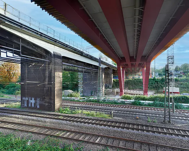 Die Unterseite einer Brücke auf roten V-Förmigen Stahlpfeilern über Bahngleisen.
