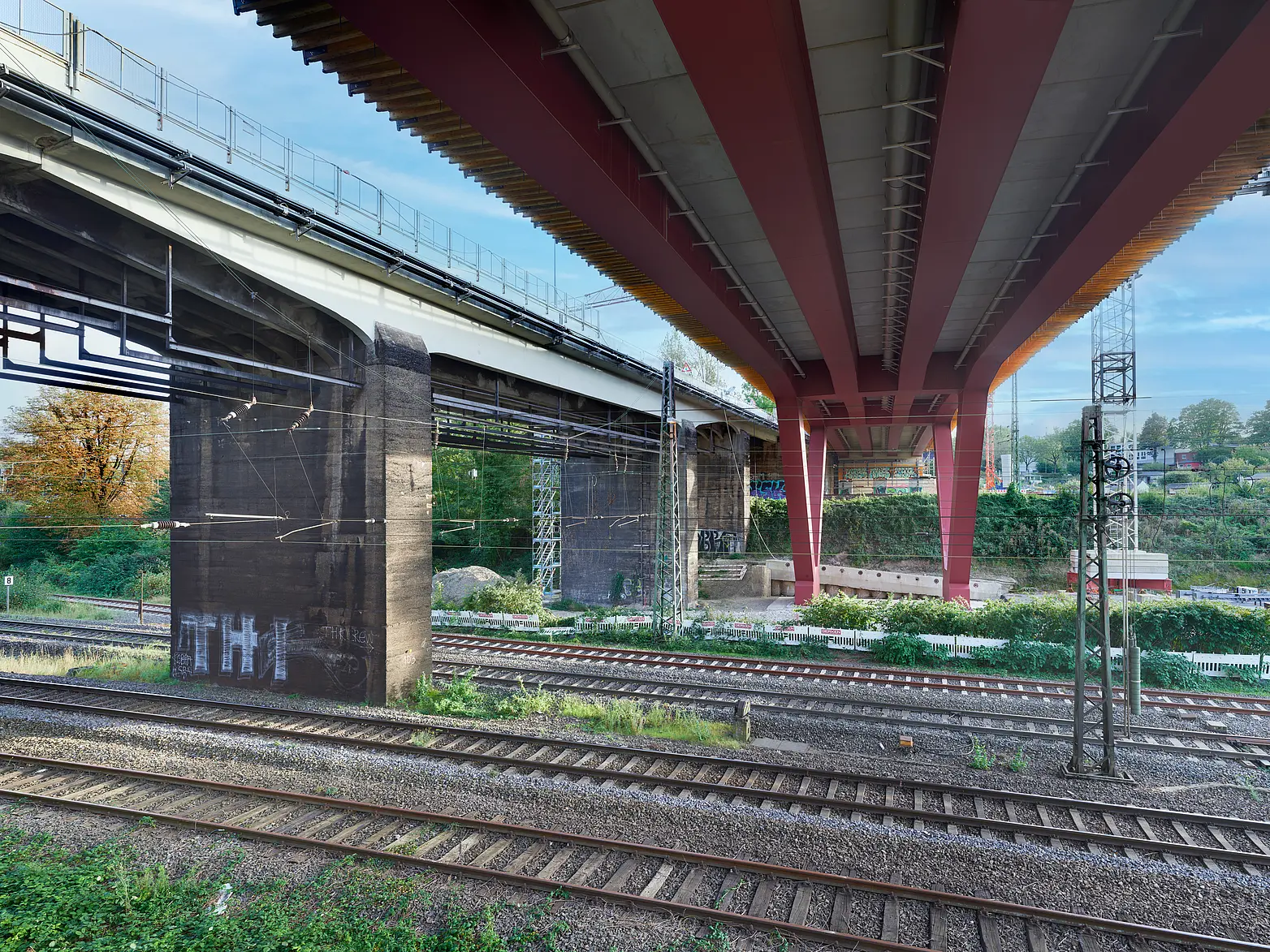 Die Unterseite einer Brücke auf roten V-Förmigen Stahlpfeilern über Bahngleisen.