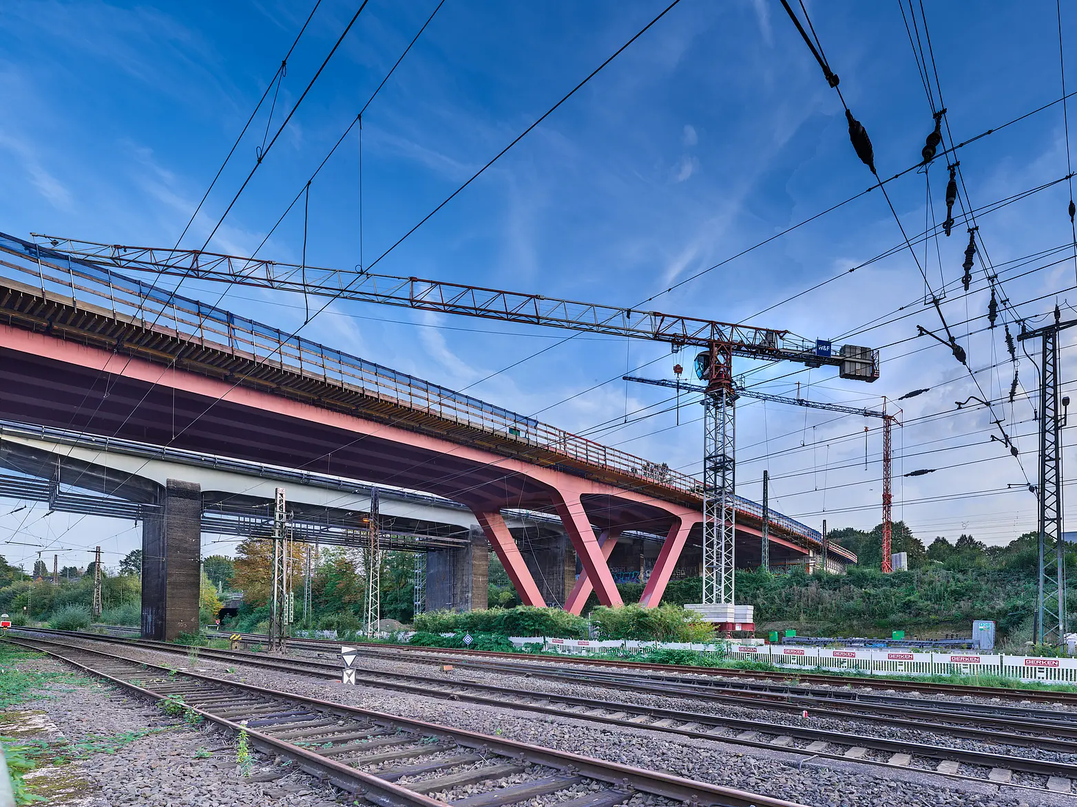 Eine Brücke auf roten V-Förmigen Stahlpfeilern über Bahngleisen, im Hintergrund ist ein Kran zu sehen.