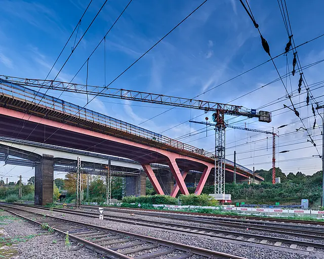 Eine Brücke auf roten V-Förmigen Stahlpfeilern über Bahngleisen, im Hintergrund ist ein Kran zu sehen.