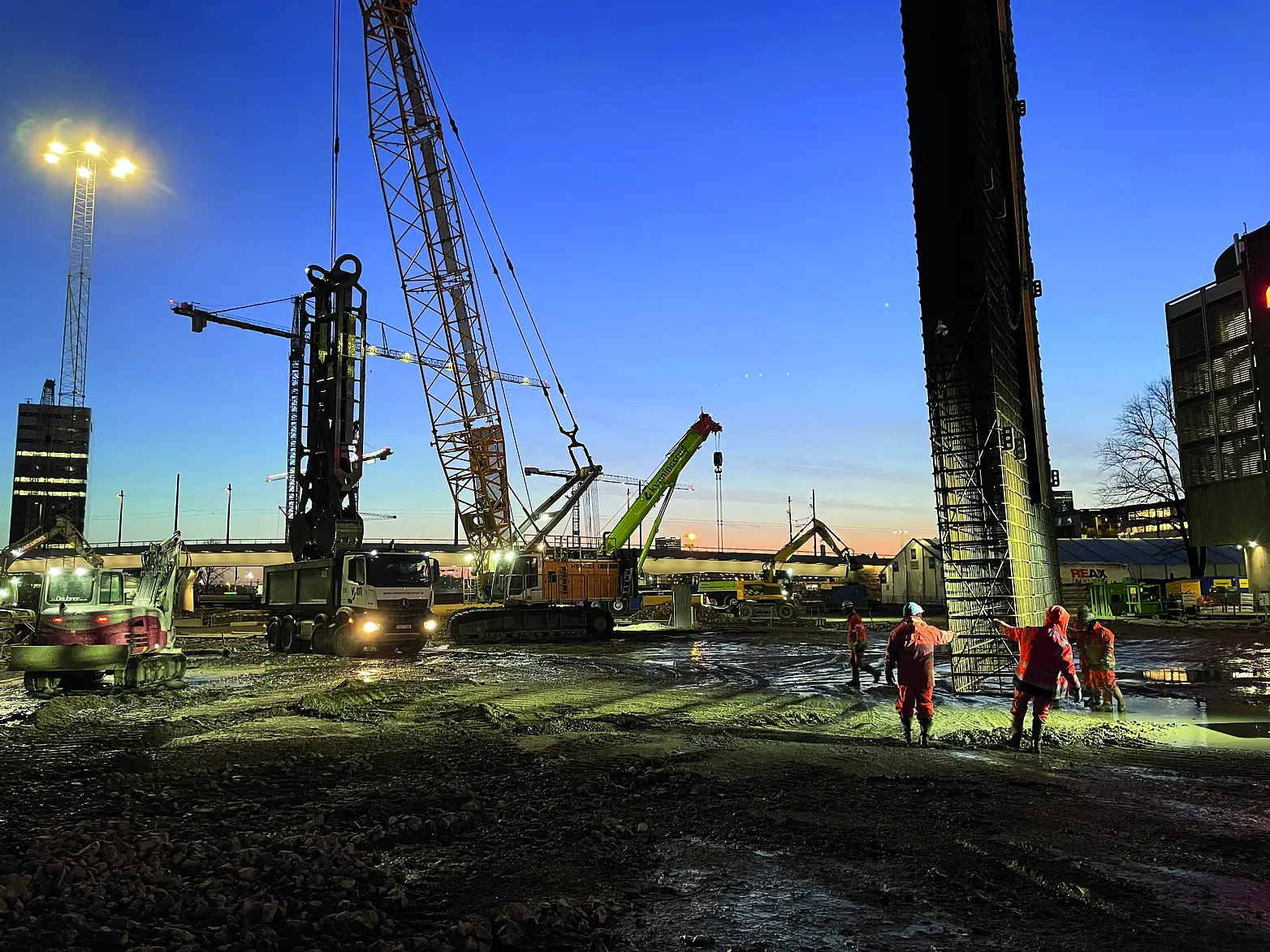 Eine Baustelle auf der Kräne und Schlitzwandgreifer zu sehen sind, die Baustelle ist liegt vor einem dunkelblauen Himmel