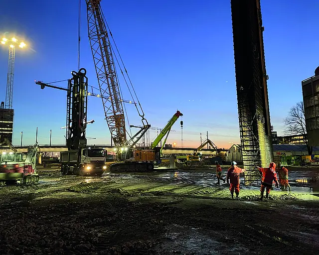 Eine Baustelle auf der Kräne und Schlitzwandgreifer zu sehen sind, die Baustelle ist liegt vor einem dunkelblauen Himmel