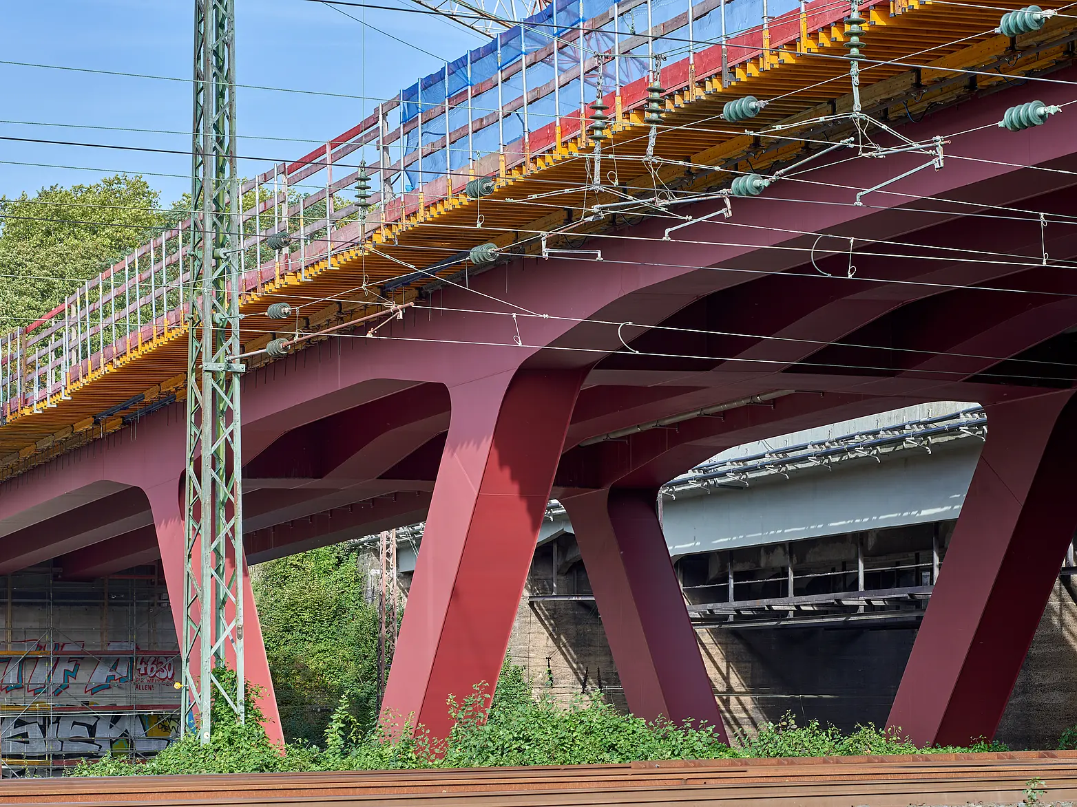 Eine Brücke auf roten V-Förmigen Stahlpfeilern über Bahngleisen.