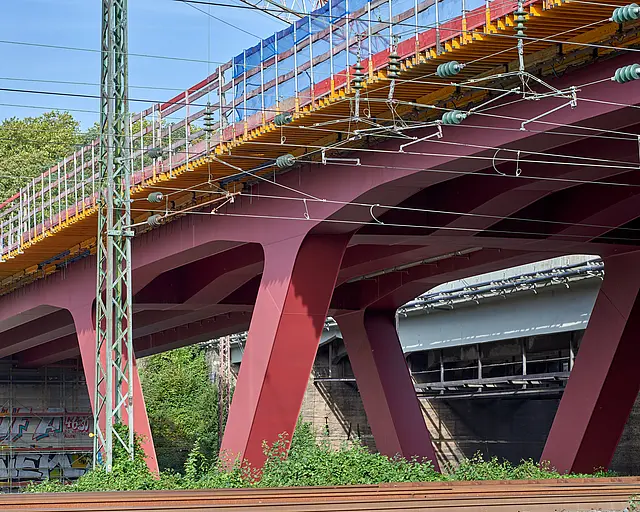 Eine Brücke auf roten V-Förmigen Stahlpfeilern über Bahngleisen.