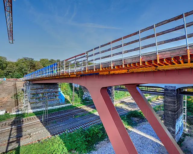 Eine Brücke auf roten V-Förmigen Stahlpfeilern über Bahngleisen, der Teil eines Krans ist zu sehen.