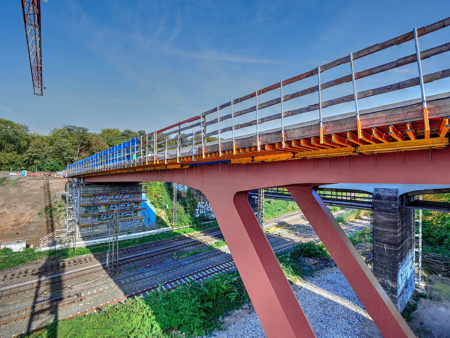 Eine Brücke auf roten V-Förmigen Stahlpfeilern über Bahngleisen, der Teil eines Krans ist zu sehen.